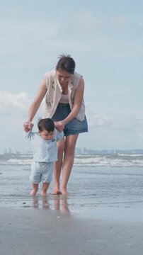 4K Asian mother holding her baby boy's hands, supporting his first steps on the beach.	
