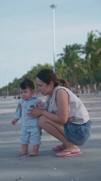 4K Asian mother holding her baby boy's hands, supporting his first steps on the beach.	
