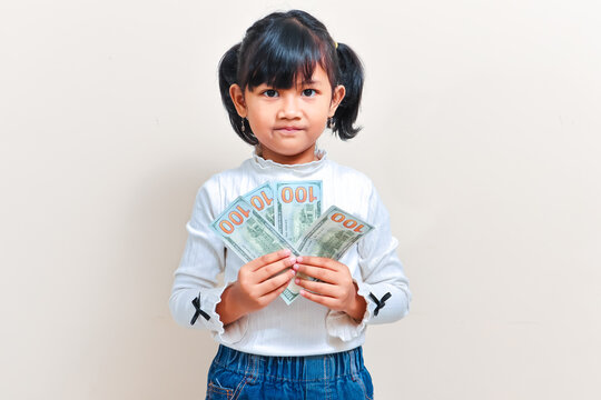 A cute little Asian girl with pigtails hair smiling while holding fan of one hundred US dollar banknotes against an isolated clean background.