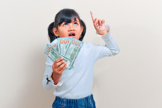 A cute little Asian girl with pigtails hair holding fan of banknotes while pointing her finger up with a thinking expression against an isolated clean background.