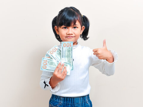 A cute little Asian girl with pigtails hair smiling cheerfully while holding fan banknotes and giving thumbs up gesture against an isolated clean background.
