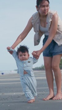 4K Asian mother holding her baby boy's hands, supporting his first steps on the beach.	
