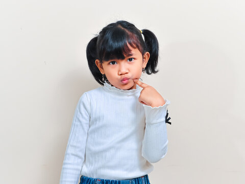 A cute little Asian girl with pigtails hair looking at the camera with a confused expression while pointing a finger at her cheek against an isolated background.