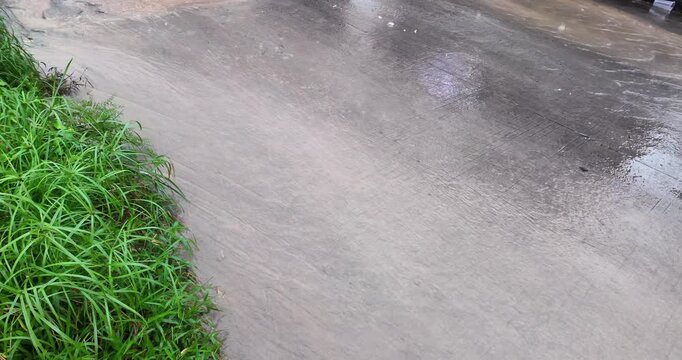 Slow motion video of rainwater flowing down a sloping road during heavy rainfall, moving past a patch of vibrant green grass at the edge.