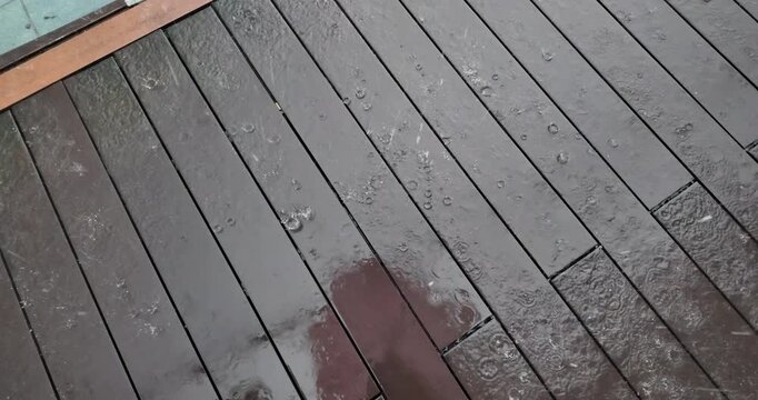 Slow motion video of raindrops creating ripples and splashes on dark, wet wooden deck planks during a rainstorm.