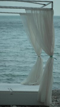 Beach canopy ocean, white bed with flowing curtains by calm sea, serene vacation spot. Vertical video.