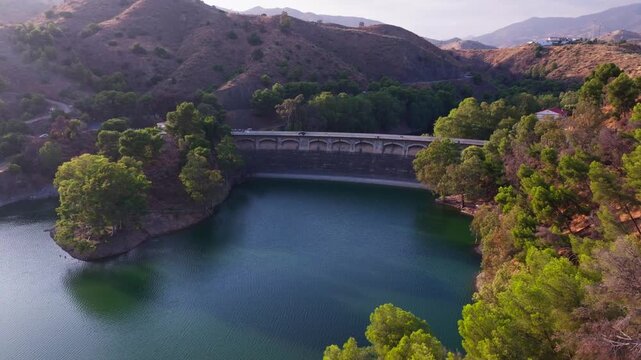 Drone footage flying forward toward the Embalse del Limonero dam near Malaga, Spain, passing a car crossing the arched bridge above teal waters.