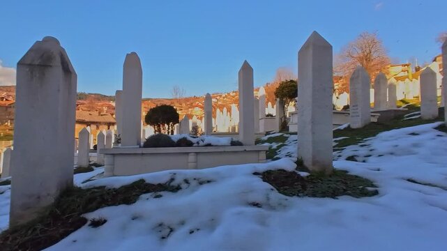 Slow panoramic panning shot of Kovači Martyr Cemetery in Sarajevo, Bosnia and Herzegovina. The rows of traditional Islamic white marble tombstones (Nisani) stand on a hillside covered with snow.
