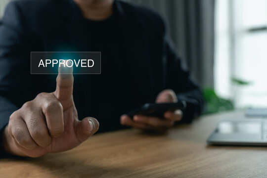 Businessman pressing an approved button on a virtual screen while holding a smartphone at his desk, concept of validation, quality assurance, and business agreement