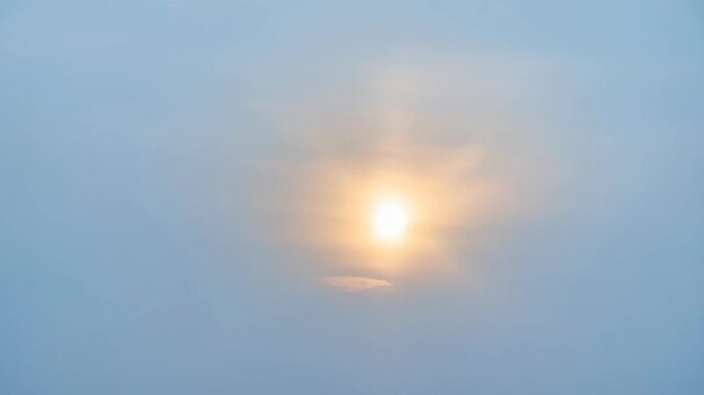 Low angle timelapse sun on blue sky concept. Bright glowing sun shining through soft blue misty sky with gentle clouds
