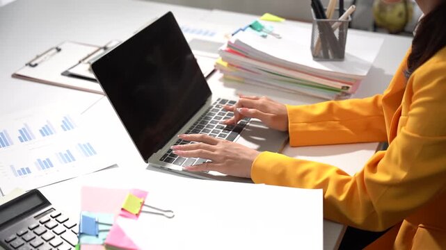 Focused Workstation: A woman's hands typing on a laptop amidst a cluttered but organized desk with charts, papers, and office supplies, symbolizing productivity and dedication.