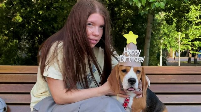 Young woman celebrates her beagle dog's birthday on a park bench outdoors, pet party, joyful companionship, animal lover lifestyle, festive moment.