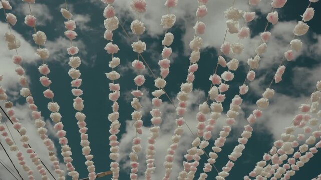 Rows of handmade paper flower garlands hang overhead against blue sky and clouds during arraial festival
