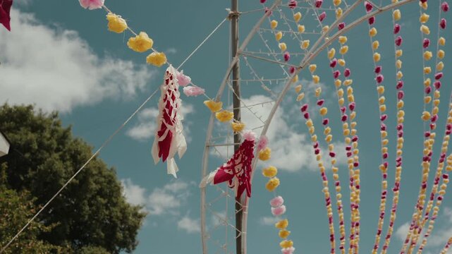 Red streamers and colorful pompom strings hang across a metal pylon against sky at arraial festival