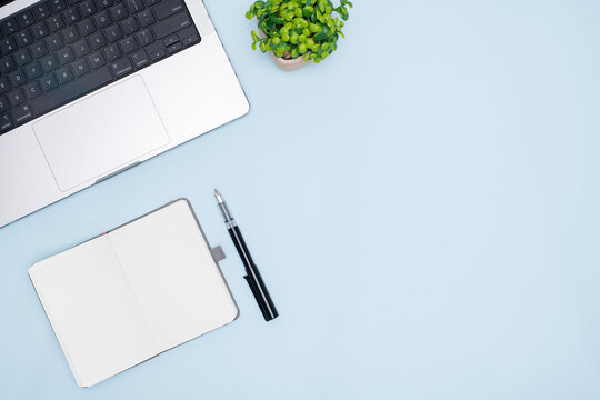 Top View of Office Workspace with Laptop, Open Notebook, Pen, and Plant on Pastel Blue Background