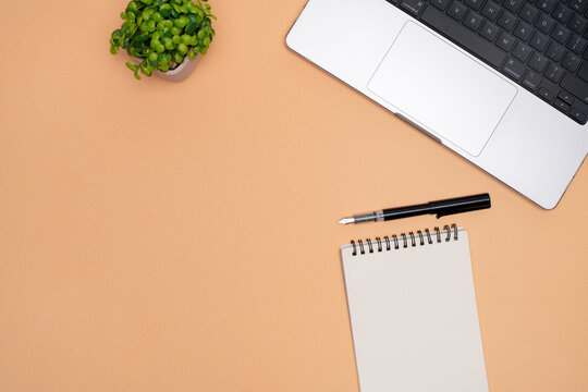 Top View of Office Workspace with Laptop, Spiral Notebook, Fountain Pen, and Plant on Peach Background