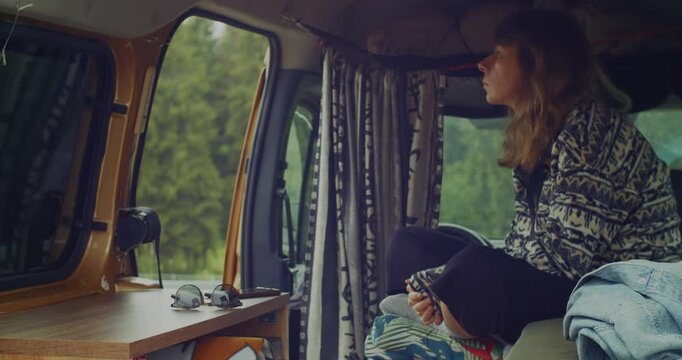 A young woman sits comfortably inside her camper van, gazing thoughtfully out the window at a lush green forest, embodying a sense of freedom and wanderlust.