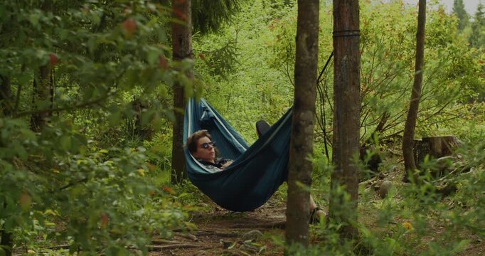 A man in sunglasses enjoys a peaceful nap in a blue hammock suspended between trees in a vibrant, ecofriendly forest setting.