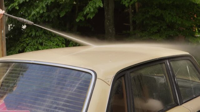 High pressure spray moves over the roof of a vintage beige car beside green trees, rinsing off grime in a driveway wash.