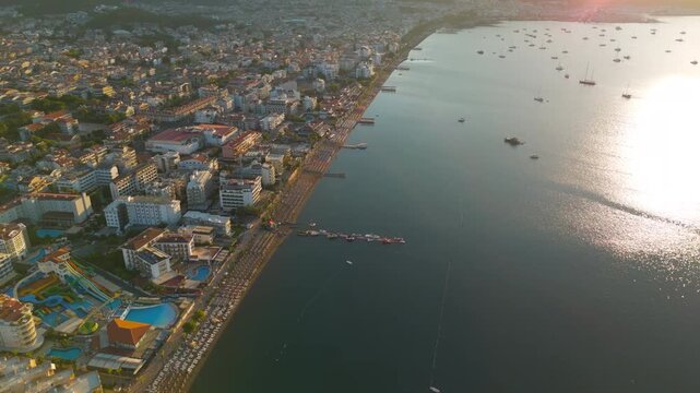 Marmaris, Turkey. Sweeping aerial view over the coastal resort strip, empty beach with rows of sunbeds and packed umbrellas awaiting tourists, hotels lining the shore in warm golden morning light. Ae