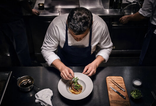 Male chef preparing delicious fresh food on ceramic plate in clean kitchen interior, side view of professional culinary expert cooking gourmet dish in restaurant