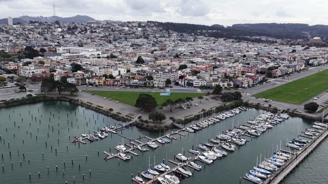 Countless sailboats pack San Francisco's Marina District harbor docks, pastel row houses and green park spread toward hazy hillside neighborhoods beneath overcast sky. Aerial drone approaching shot