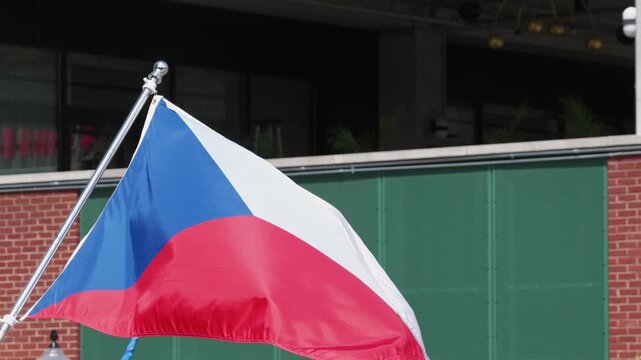 Czech Republic flag ripples atop tall pole beside weathered brick building in Lancaster, green garage door and additional international flags flutter nearby beneath bright midday sunlight.