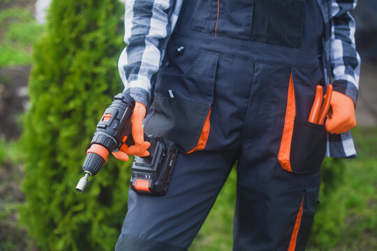 person holding power drill outdoors wearing orange gloves and black jacket for home improvement project in garden setting close-up practical and serene mood natural light clean background.