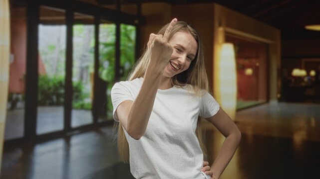 Woman smiling shows middle finger with raised hand in building lobby with glass doors and lamps visible; defiance.