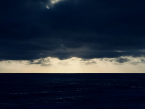 A wide dramatic landscape view of a calm dark blue sea under heavy gloomy overcast clouds on a July summer morning in Carlsbad, California.