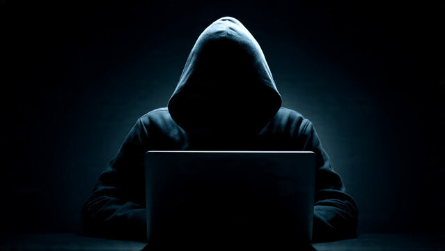 Silhouette of a hooded person using a laptop in dramatic low light, concept for cybersecurity, digital crime and data privacy awareness