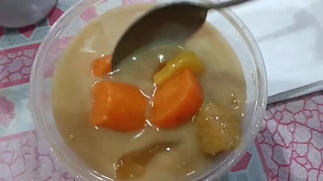 Close-up of sweet Indonesian fruit compote in a plastic cup with spoon, containing sweet potato, banana and coconut milk broth. Kolak