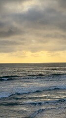 Fototapeta premium A portrait layout vertical panorama capturing layered grey storm clouds over golden light reflecting on rolling ocean swell waves in Carlsbad, California.