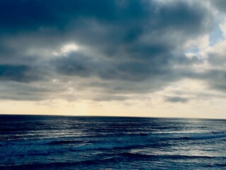 Fototapeta premium A wide horizontal layout showing heavy, dark moody overcast storm clouds hanging low above a churning textured deep blue Pacific Ocean surface in Carlsbad, California.