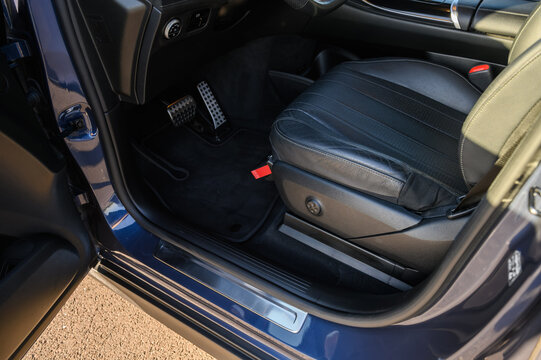 Close up of a modern luxury car driver side interior with open door, black leather seat, pedals, floor mat, and sill plate.