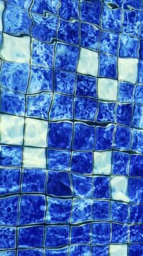 Blue mosaic swimming pool tiles shimmer beneath clear rippling water and sunlight.