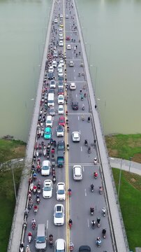 Vertical screen Aerial Hue Bridge Packed With Vehicles And Scooters During Evening Rush, Dense Queue Stretches Across River With Park And Palm Trees On Banks Slow Gridlock And Honking