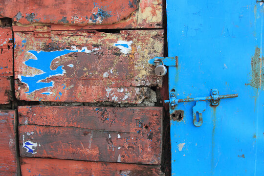 Close-up of a weathered red metal door secured with a padlock, featuring peeling paint and a rustic textured surface
