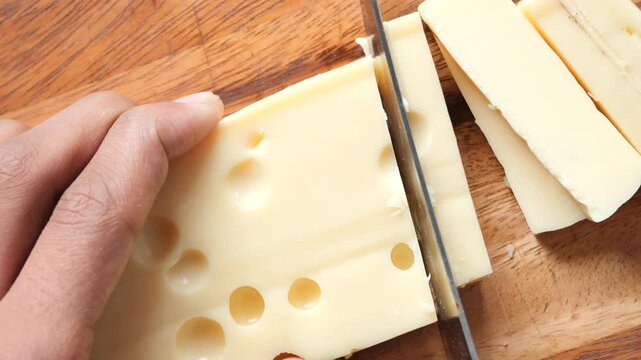 Close up slow motion video footage of hand slicing delicious yellow cheddar cheese block with knife on wooden cutting board