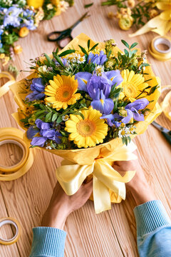 A person holds a bouquet of yellow and purple flowers tied with a ribbon. The wooden table has scissors and wrapping materials scattered around