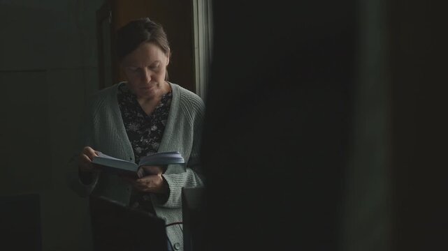 Female figure in low-lit old office room holding open book by the window, calm reading scene with moody atmosphere, 4K