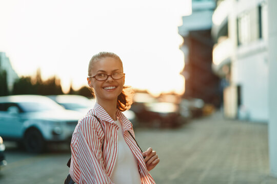 A smiling young woman in striped shirt with glasses walks along a sunny urban street, radiating confidence and calm, ready for a casual day.