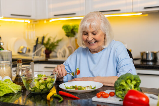 Senior old woman smiling while eating fresh vegetable salad at home kitchen for diet support. Elderly grandmother enjoying healthy safe meal choice with calm mood for wellness nutrition goal indoors.
