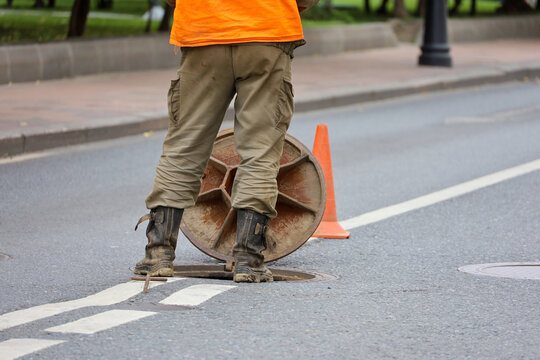 Worker in uniform standing over the open sewer hatch on city street. Concept of repair of sewage, underground utilities, water supply system, cable laying, water pipe accident