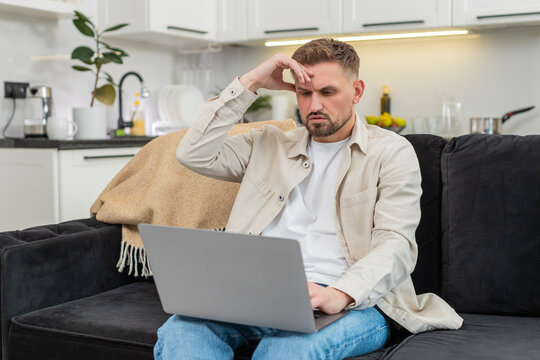Young man at home looks at laptop sees defeat touches forehead sighs and clicks retry button. Caucasian guy on sofa watches screen shows frustration sad mood after bad news and failed attempt again.