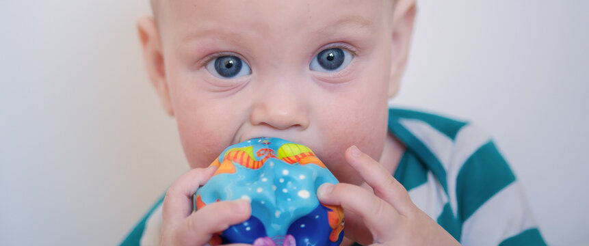 Tiny child with sparkling blue eyes focuses intently while gnawing on a bright patterned textured toy during a peaceful moment of discovery in a sunlit indoor home environment