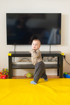 Little child stands on a bright yellow mat while exploring the world, holding onto furniture with tiny hands during a quiet morning spent indoors at home