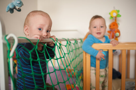 Twin babies hold onto the colorful safety netting of their small wooden beds while interacting and smiling in a warm home environment during the daytime