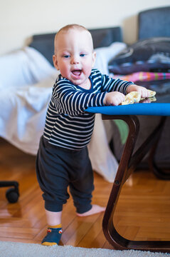 Joyful baby boy in a striped shirt stands confidently holding onto a low table during a bright morning at home while learning to balance on his feet