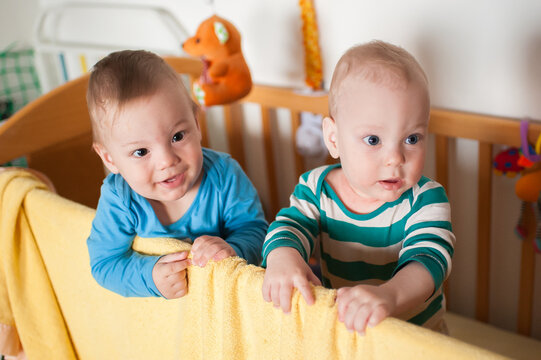 Sweet twin brothers discover the world from the safety of their wooden nursery bed, gazing ahead with wonder and delight as they hold onto the soft yellow blanket during a quiet start to the day